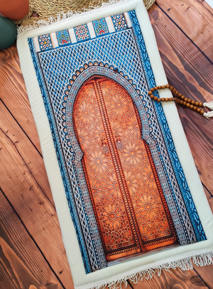 A rectangular prayer rug laid flat on a wooden floor, featuring a printed arched door design in warm orange-brown tones surrounded by blue and white ornamental borders, with white fringe along the edges. A string of wooden prayer beads rests beside the rug.