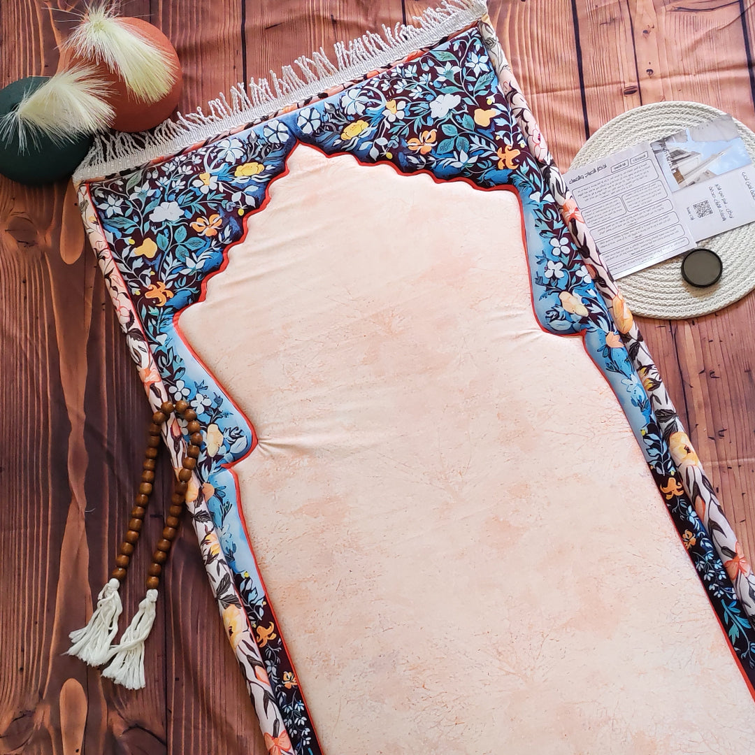A decorative prayer rug laid flat on a wooden surface, featuring a large pale blush center, a multicolor floral border with blue, orange, and cream tones, fringe along the top edge, and wooden prayer beads hanging from one corner. A woven mat and small decorative items are partially visible around it.